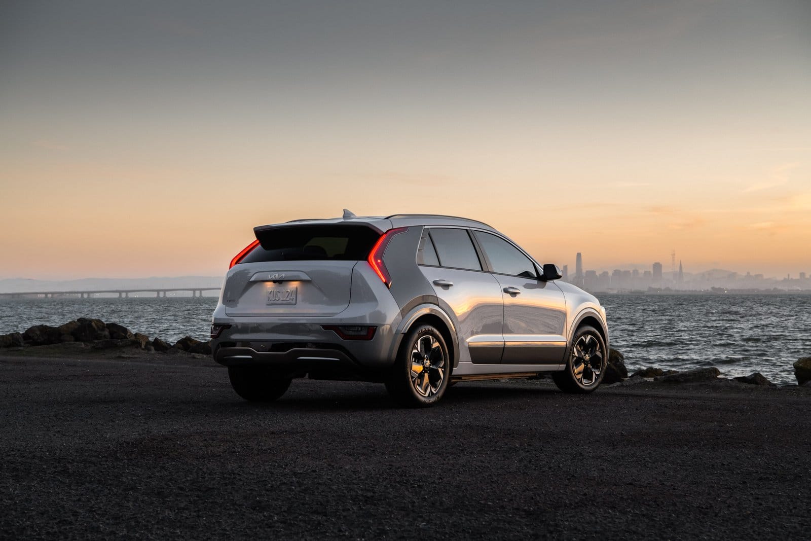 Rear and side view of a silver Kia Niro EV electric vehicle parked near the ocean at sunset with city skyline in the background