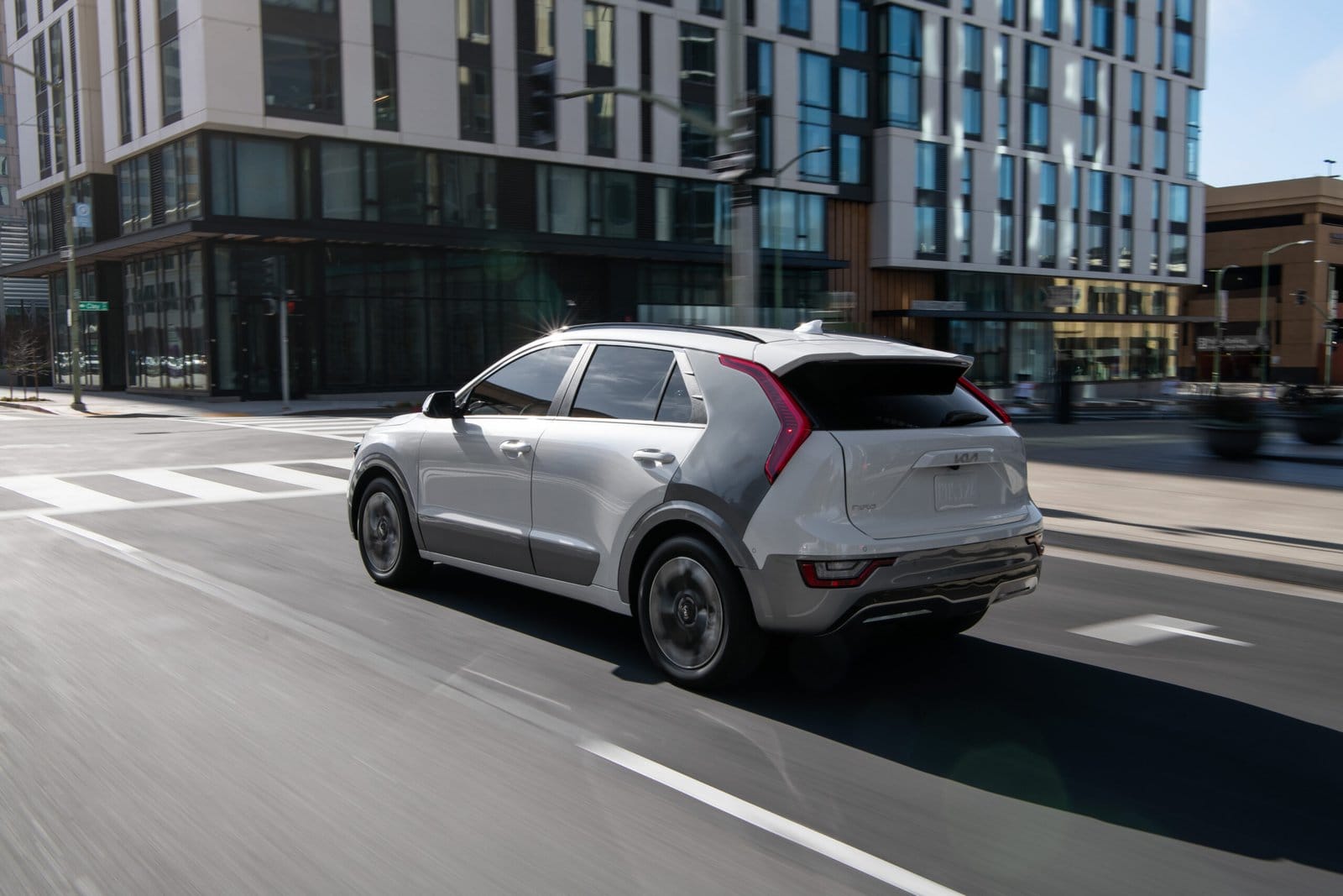 Rear side view of a Kia Niro EV electric vehicle in motion through an urban intersection with modern buildings