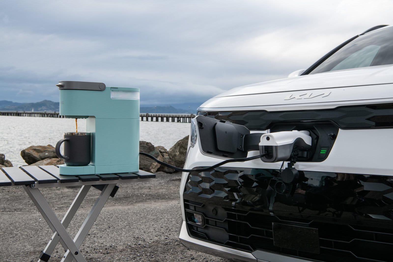 Kia Niro EV electric vehicle charging port powering a coffee maker through vehicle-to-load (V2L) on a coastal road