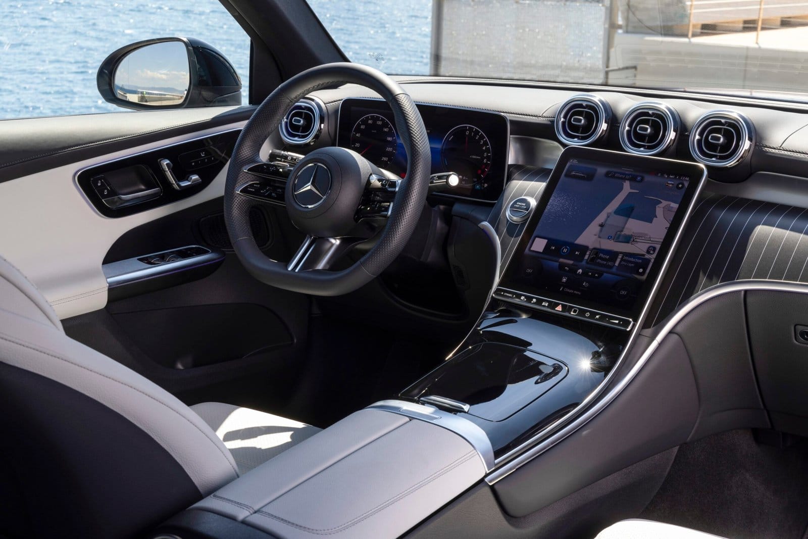 Interior of 2025 Mercedes-Benz GLC 350e electric vehicle showing steering wheel, digital dashboard, and central touchscreen with ocean view through side mirror