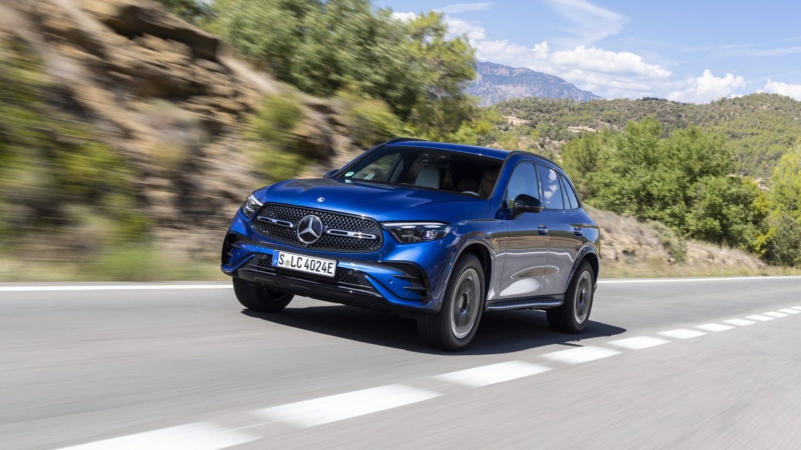 Blue 2025 Mercedes-Benz GLC 350e electric vehicle driving on a scenic road with hills and trees in the background