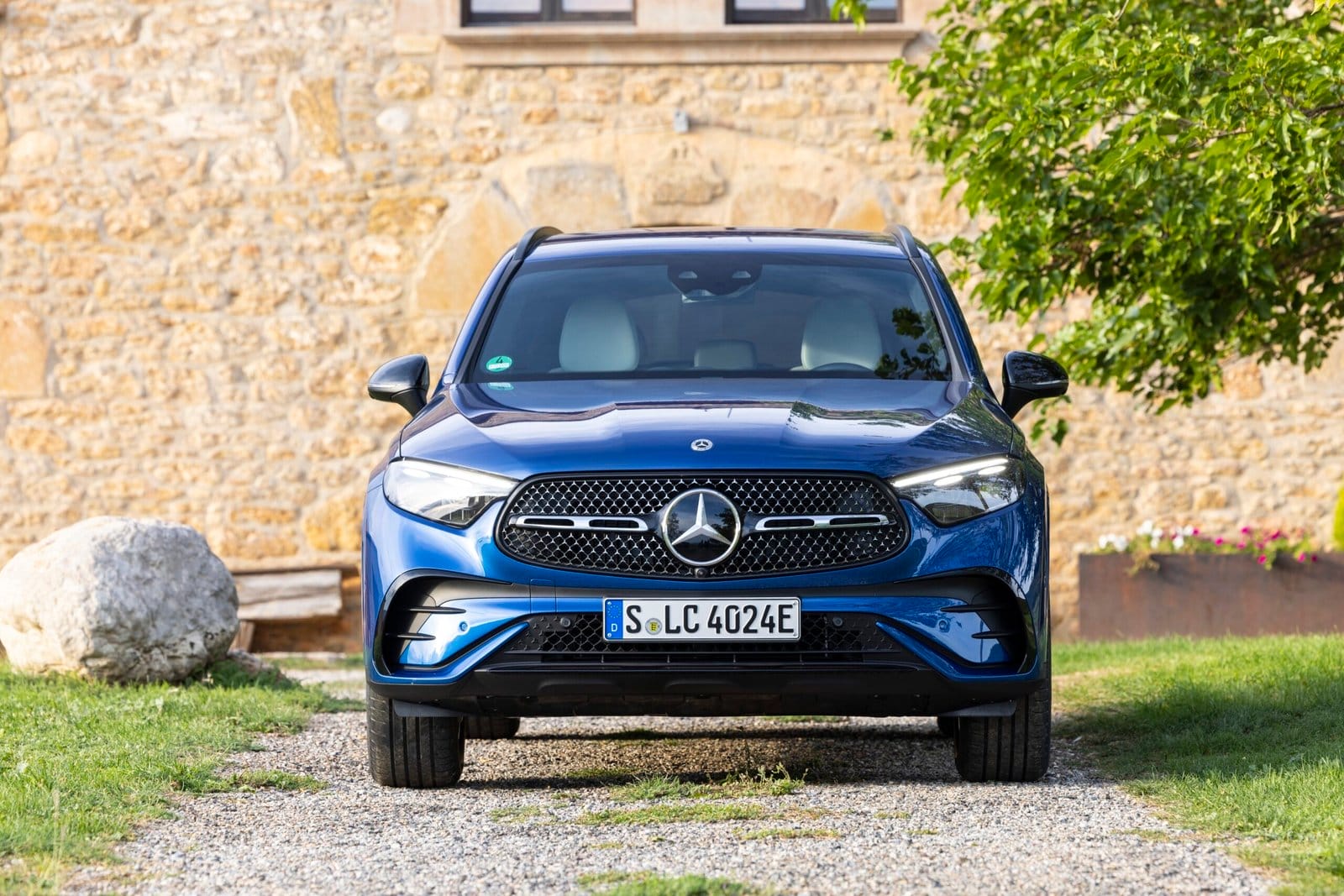 Blue 2025 Mercedes-Benz GLC 350e electric vehicle viewed from the front, parked on a gravel path near a stone wall and greenery