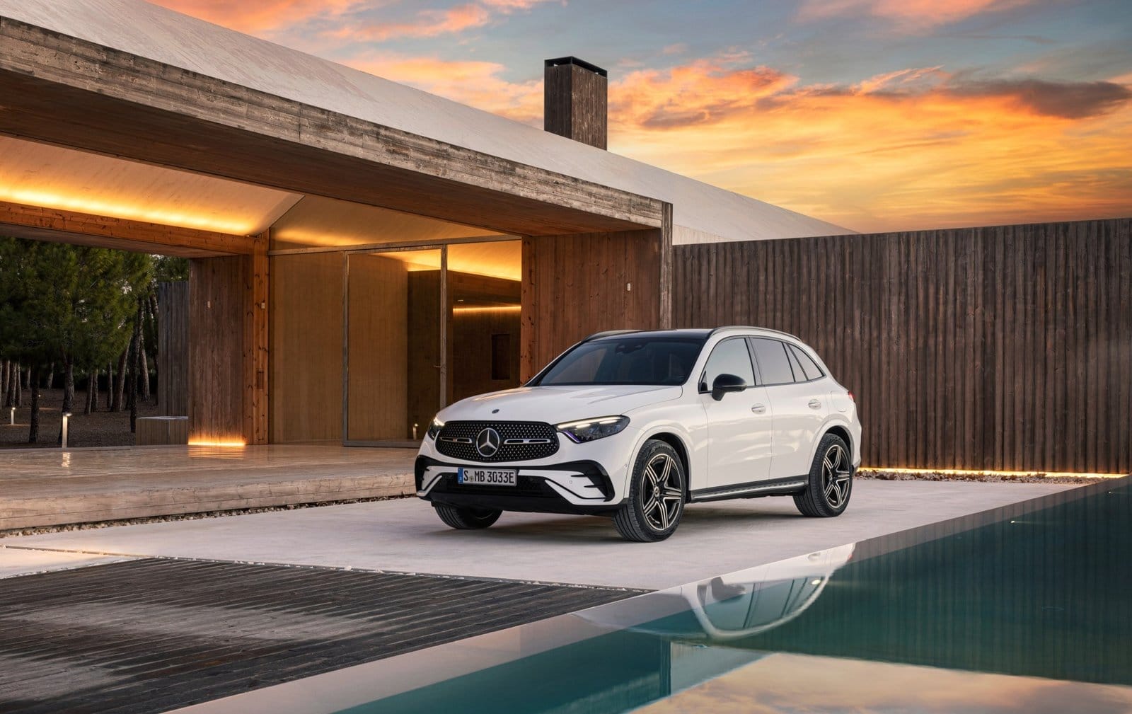 Front side view of 2025 Mercedes-Benz GLC 350e electric vehicle in white, parked beside a pool and modern wooden house at sunset