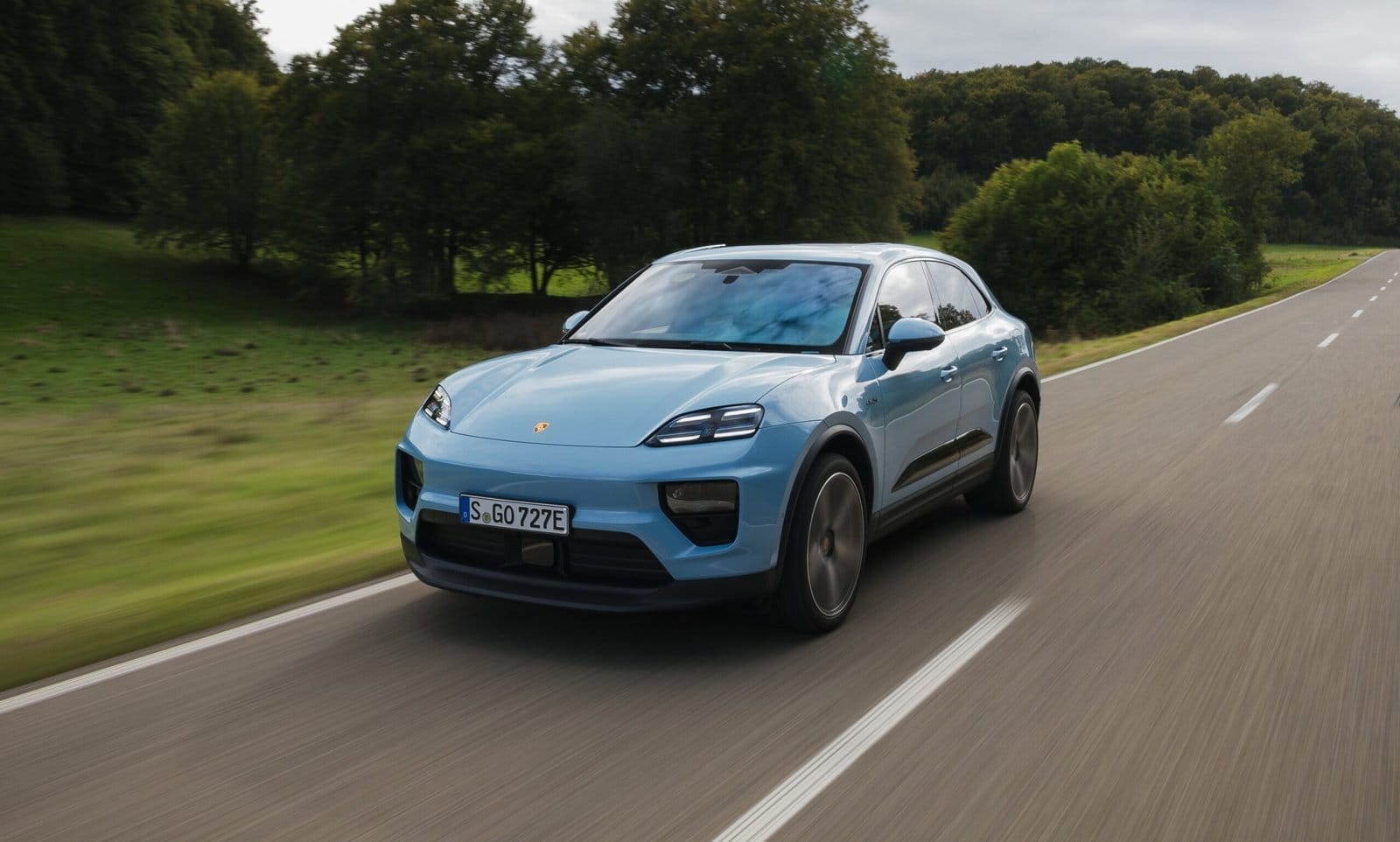 Light blue Porsche Macan EV electric vehicle captured from front-side angle while driving on a countryside road with green fields and trees