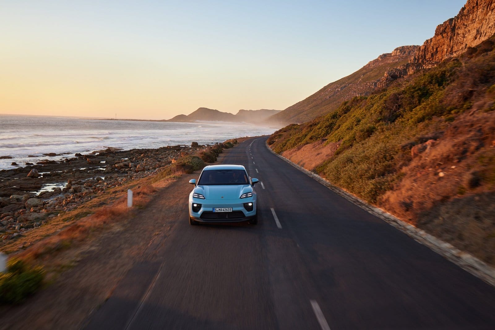 Front view of blue Porsche Macan EV electric vehicle driving along a scenic coastal road at sunset with ocean and cliffs visible