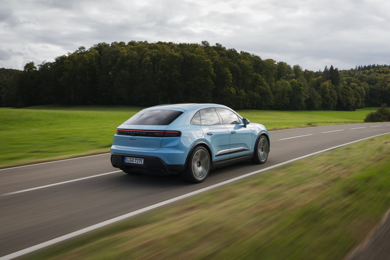 Rear-side view of blue Porsche Macan EV electric vehicle driving on a rural road surrounded by green fields and dense forest
