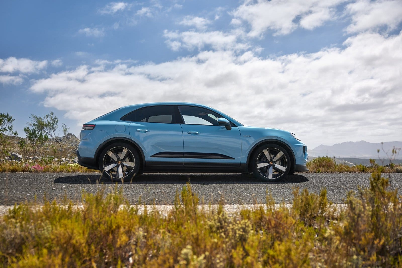 Side view of blue Porsche Macan EV electric vehicle parked on a roadside with mountains and clouds in the background