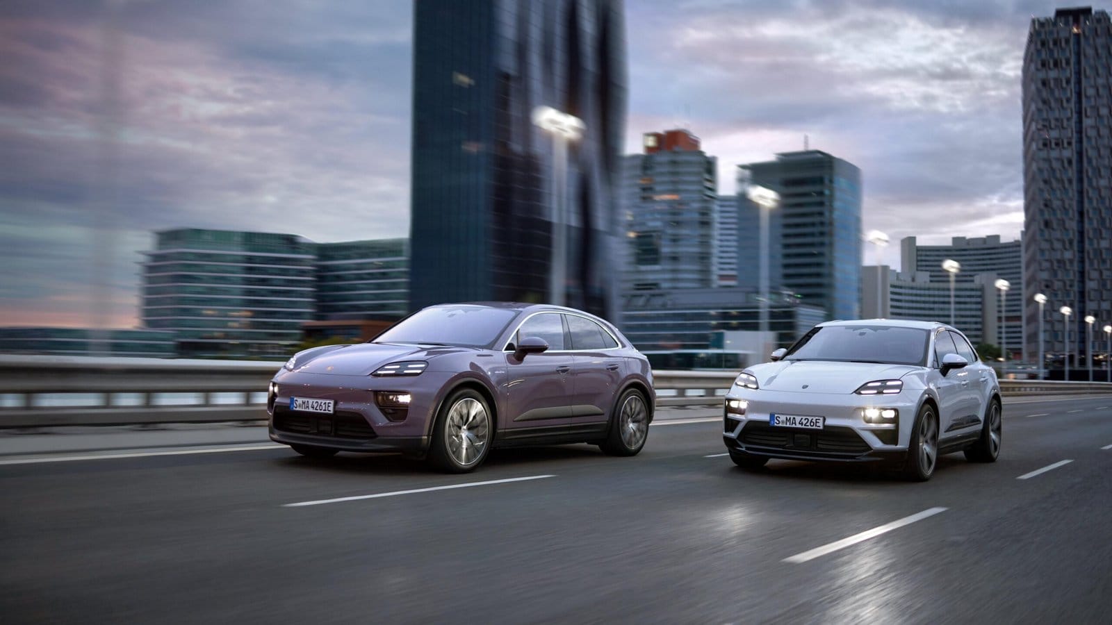 Two Porsche Macan EV electric vehicles, one white and one purple, driving side-by-side on a multi-lane road with modern high-rise buildings in the background