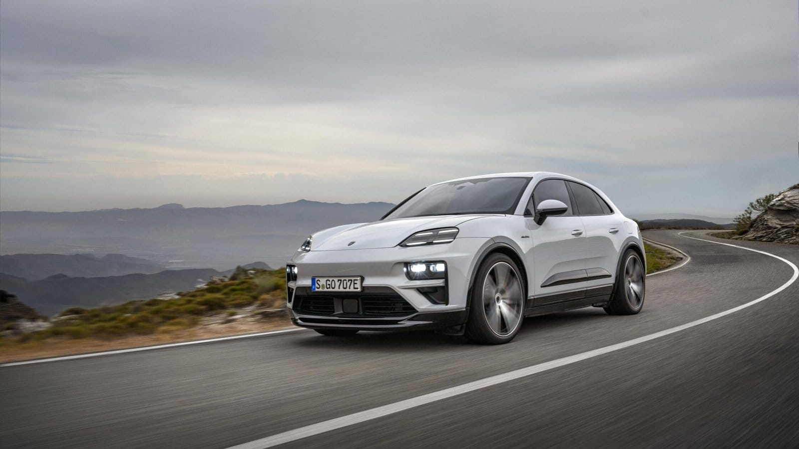 White Porsche Macan EV electric vehicle driving on a winding mountain road with cloudy sky and distant hills in the background