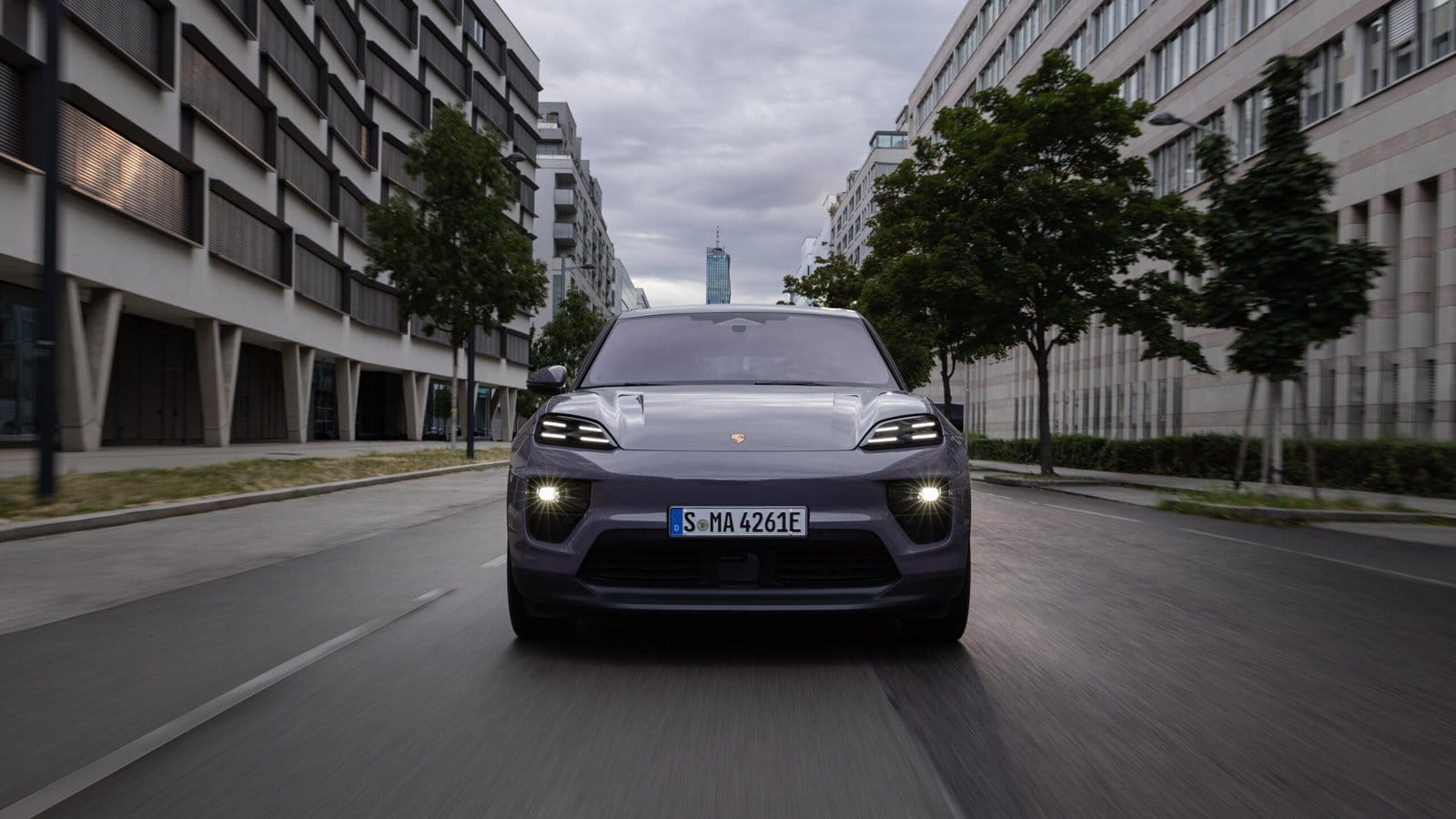 Front view of a purple Porsche Macan EV electric vehicle driving on a city street lined with modern buildings and trees