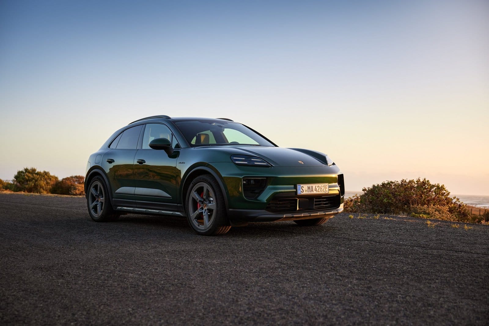 Green Porsche Macan EV electric vehicle parked on coastal road at sunset, viewed from the front side