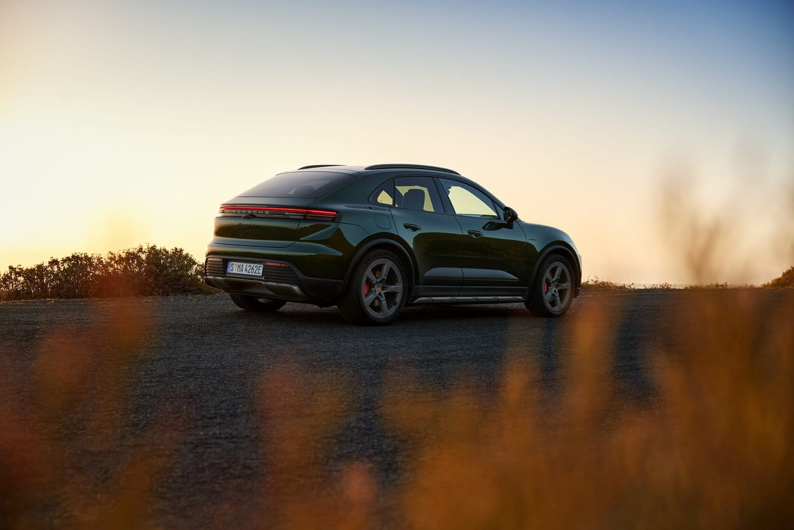 Rear-side view of green Porsche Macan EV electric vehicle parked on a scenic roadside during sunset