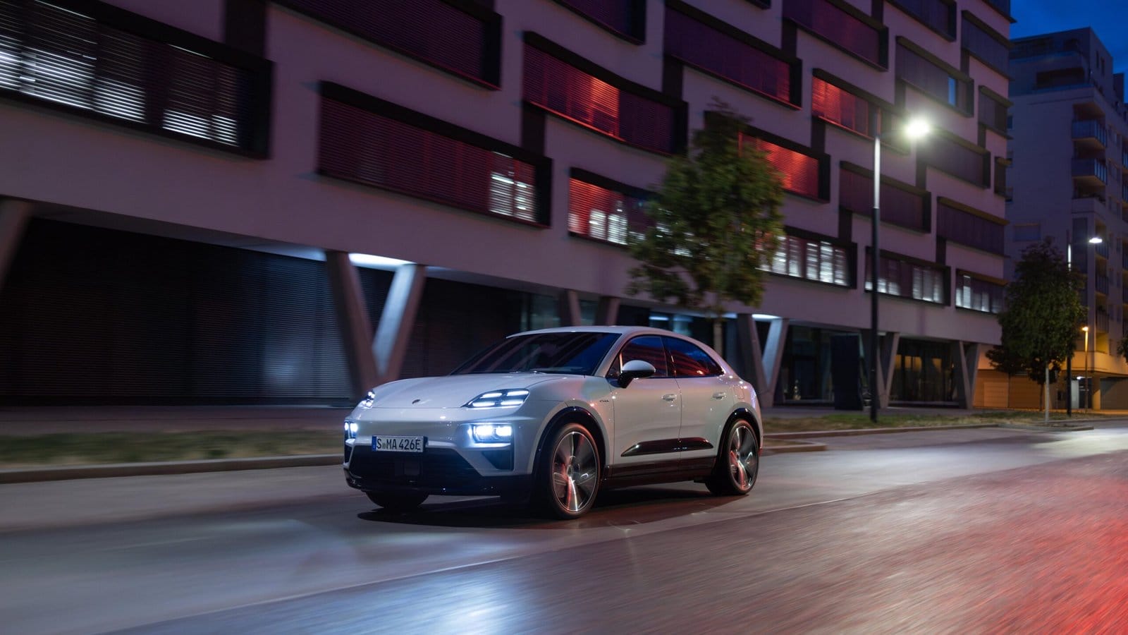 White Porsche Macan EV electric vehicle driving on a city street at night with illuminated headlights and building reflections