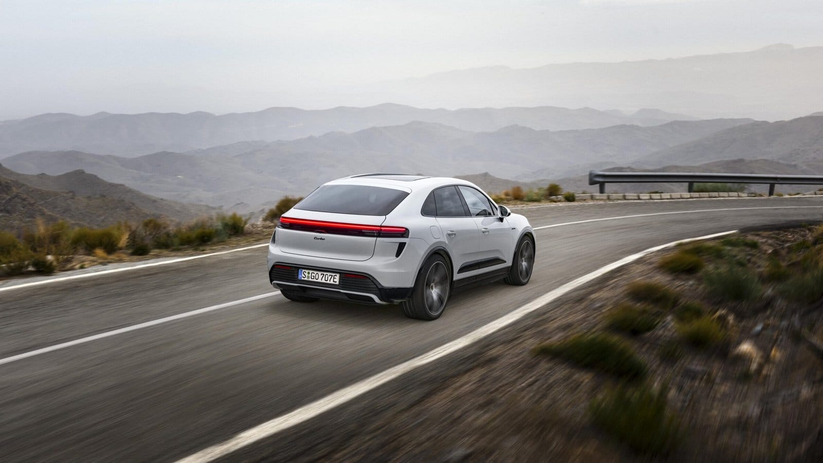 White Porsche Macan EV electric vehicle captured from rear three-quarter view while driving on a winding mountain road