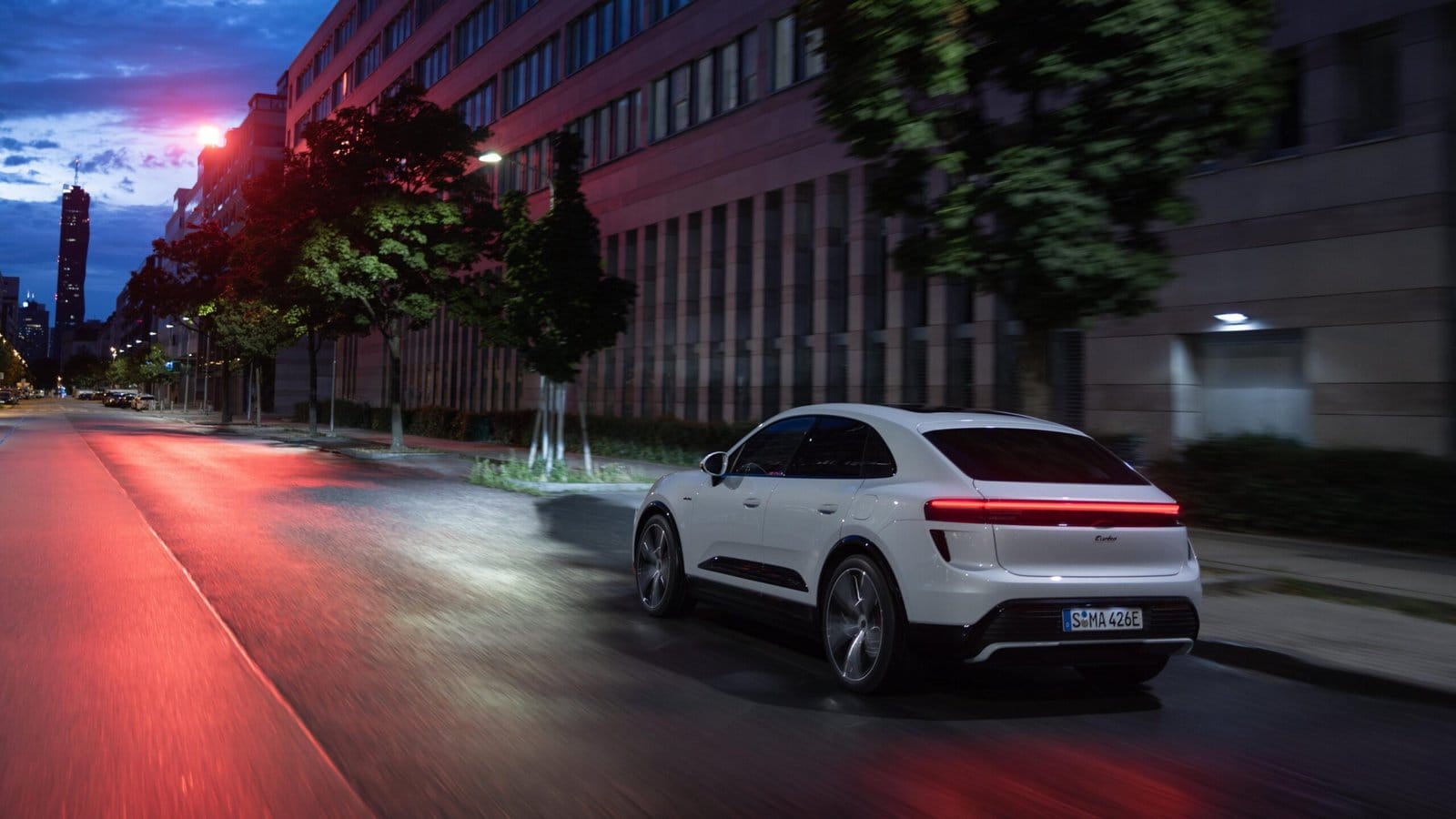 Rear view of white Porsche Macan EV electric vehicle driving through a city street at night with taillights illuminated