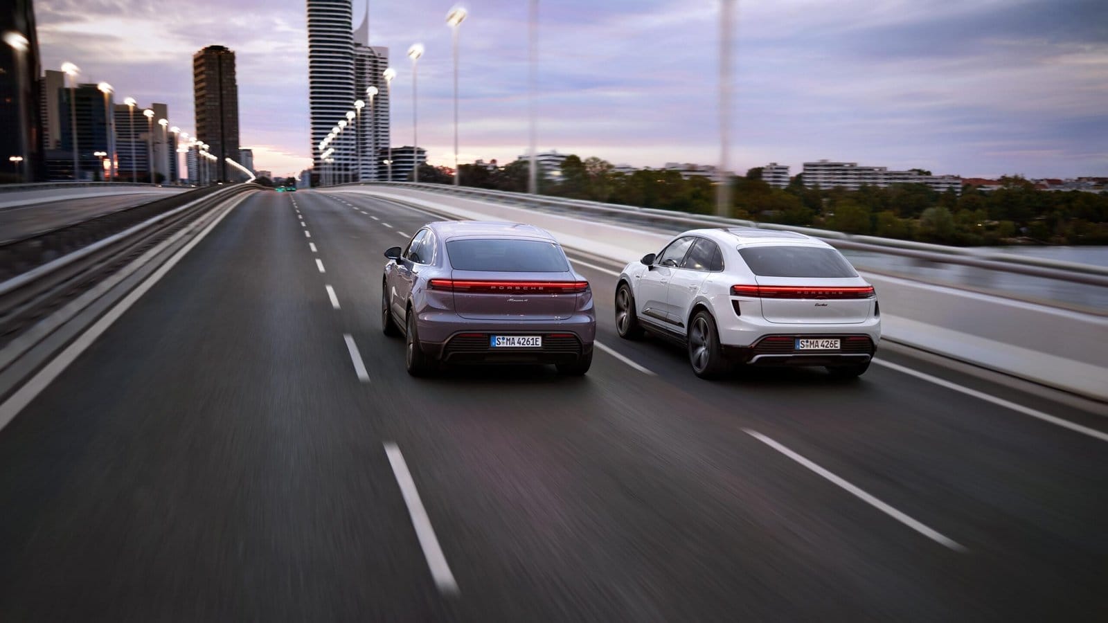 Rear view of two Porsche Macan EV electric vehicles, one in white and one in purple, driving side-by-side on a bridge with urban skyline in the background