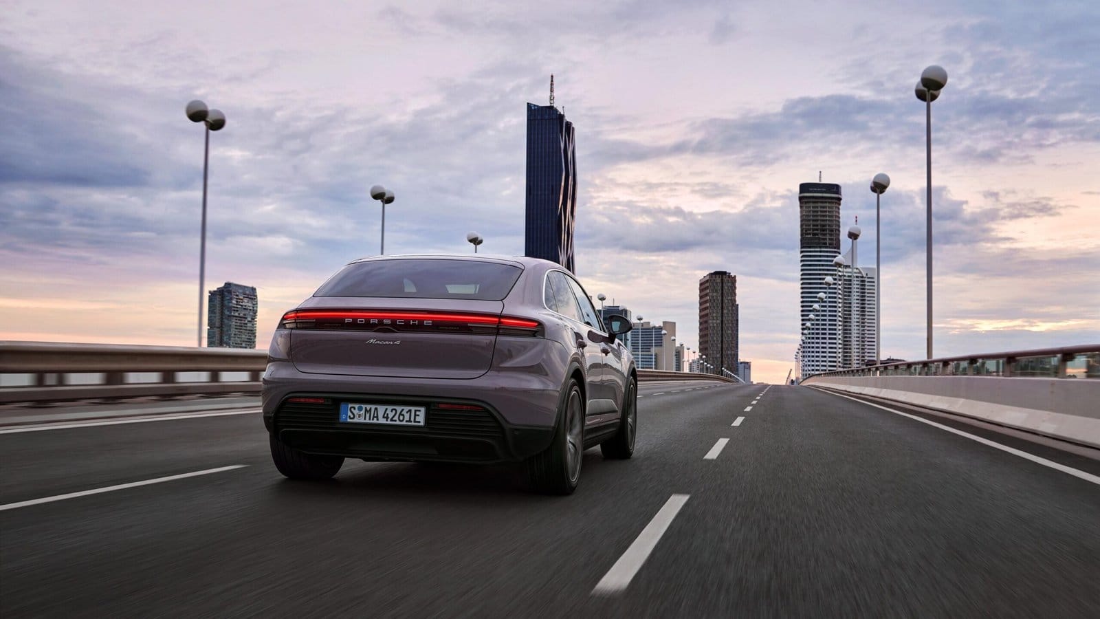 Rear view of a purple Porsche Macan EV electric vehicle driving on a bridge toward a city skyline at sunset