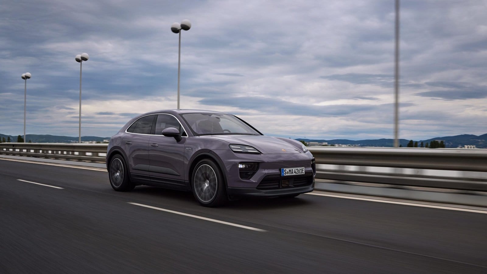 Side profile of a purple Porsche Macan EV electric vehicle in motion on a bridge with cloudy skies and guardrails in the background