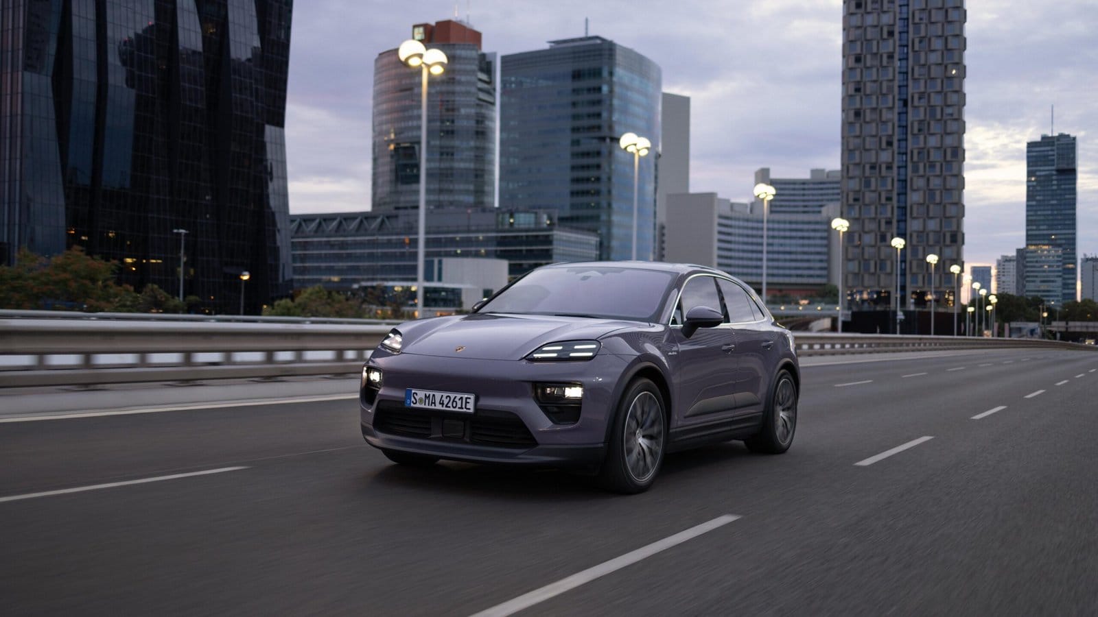 Purple Porsche Macan EV electric vehicle driving on an empty city road surrounded by modern high-rise buildings at dusk