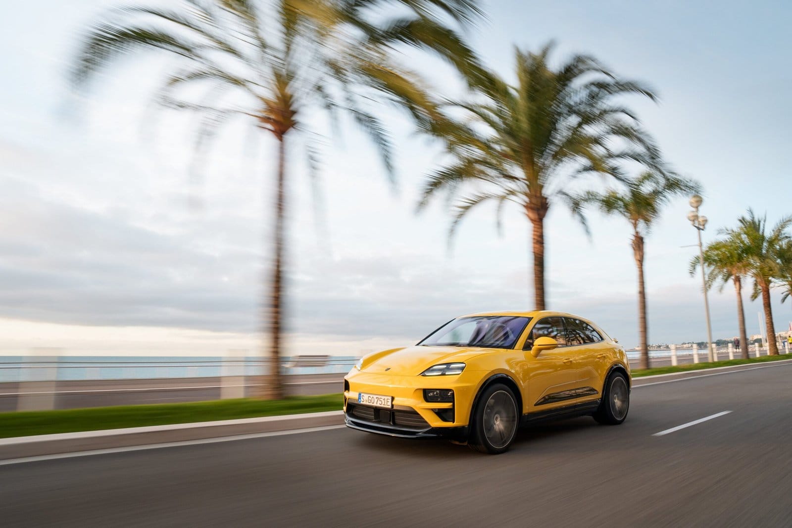 Yellow Porsche Macan EV electric vehicle captured from the front side while driving along a palm-lined road by the sea