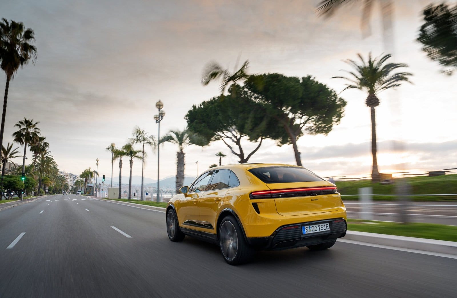 Rear-side view of yellow Porsche Macan EV electric vehicle driving on a palm-lined coastal road during sunrise