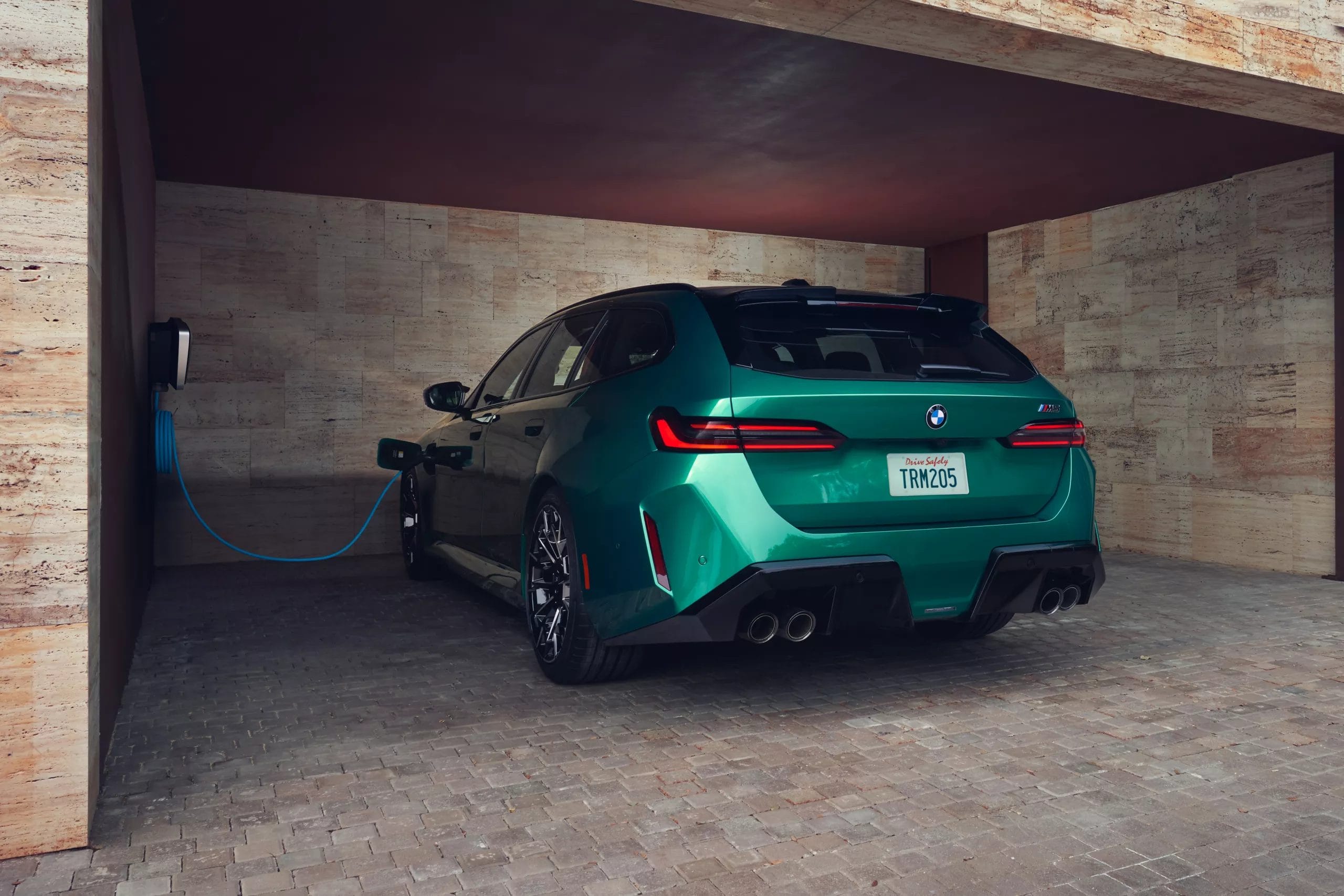 BMW M5 Touring Plug-in Hybrid in green connected to a wall-mounted charger inside a stone-walled home garage