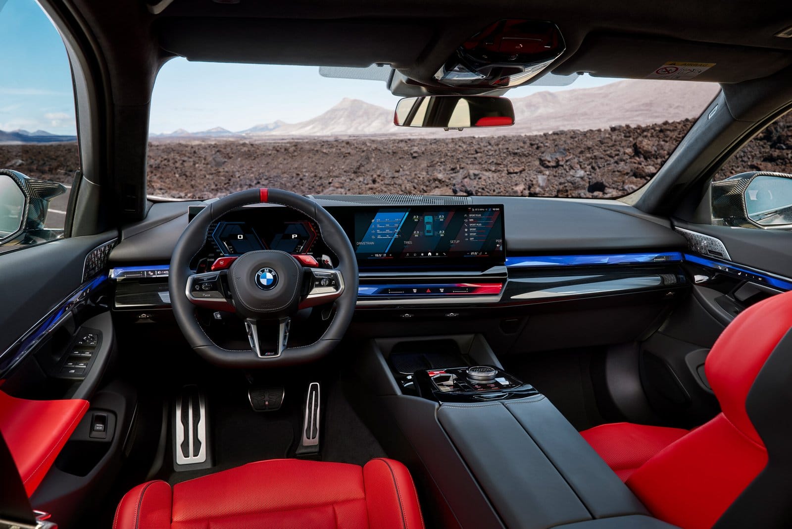 Driver's view of BMW M5 Touring Plug-in Hybrid interior with red seats, digital instrument cluster, and curved infotainment display