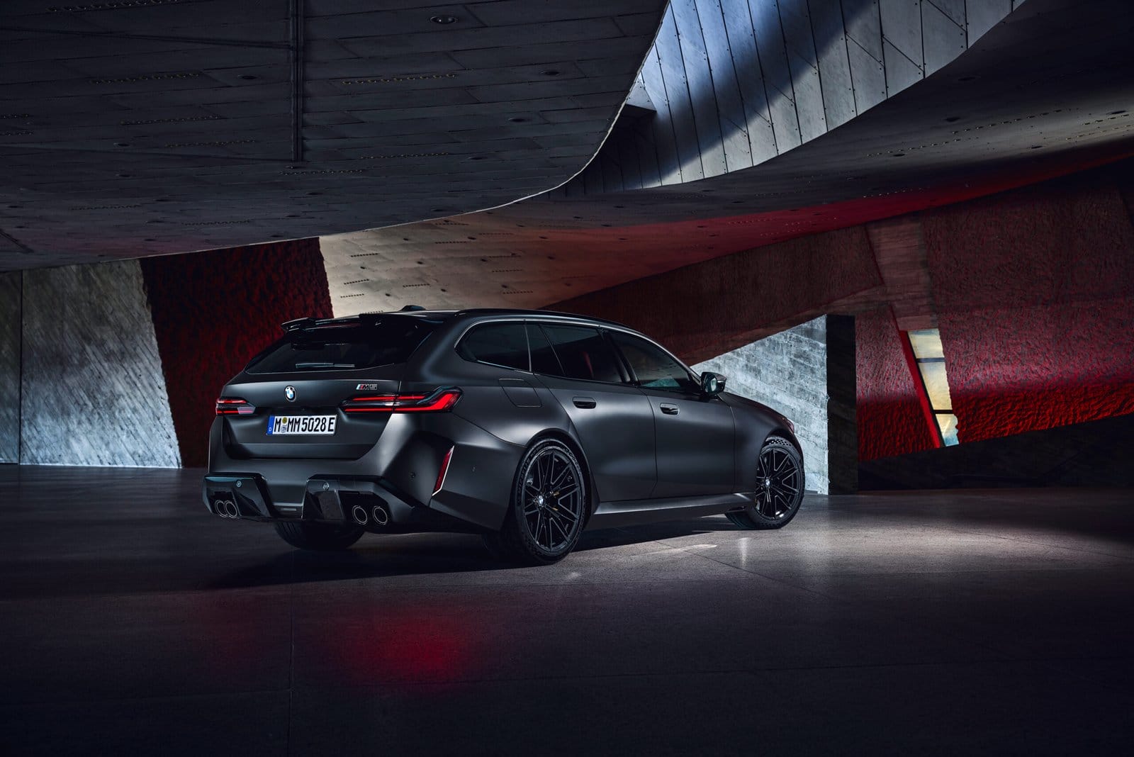 Rear view of BMW M5 Touring Plug-in Hybrid in matte black, parked under sculptural concrete and red lighting