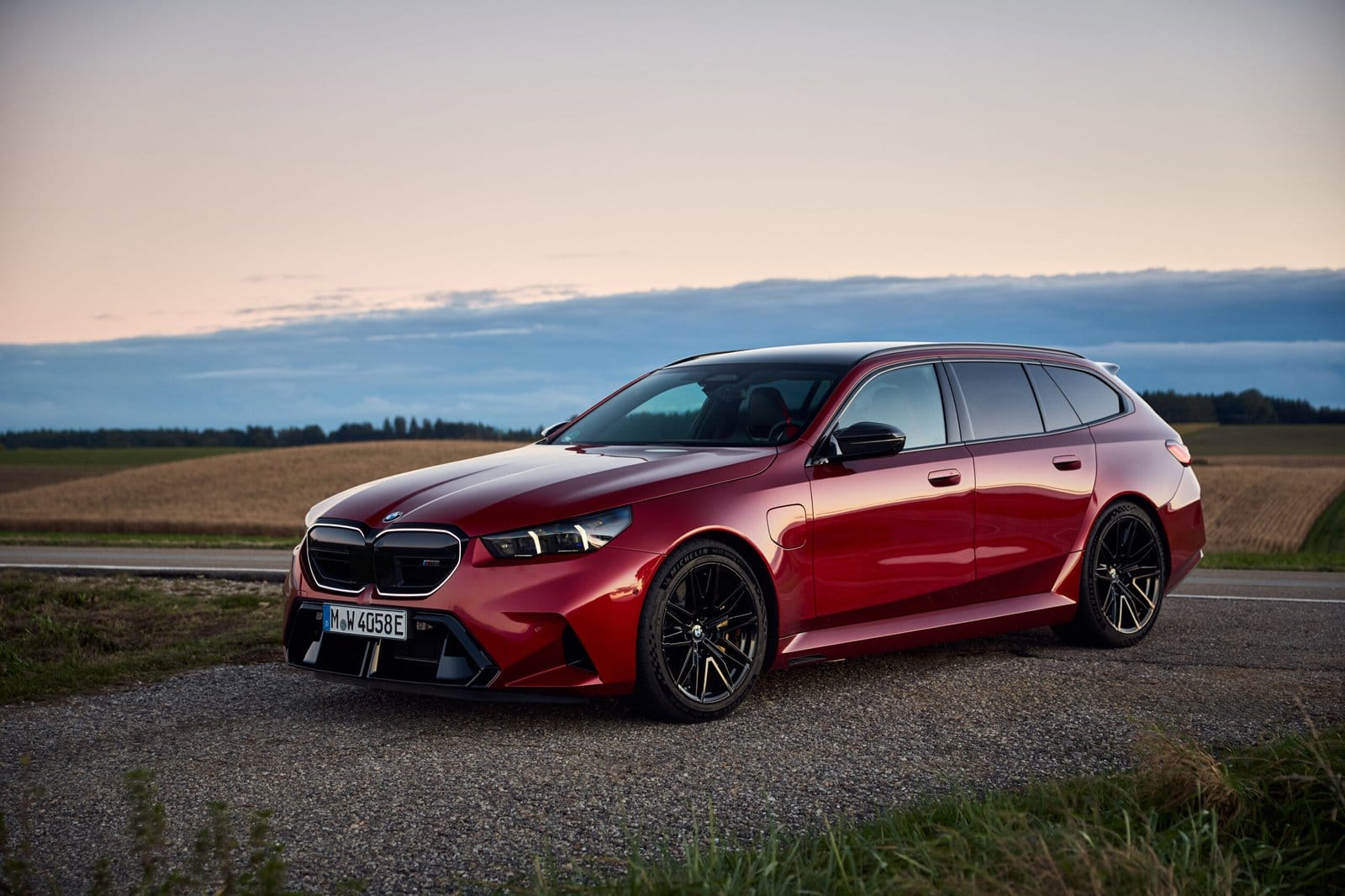 Front side view of the red 2025 BMW M5 Touring Plug-in Hybrid parked on a rural roadside at dusk