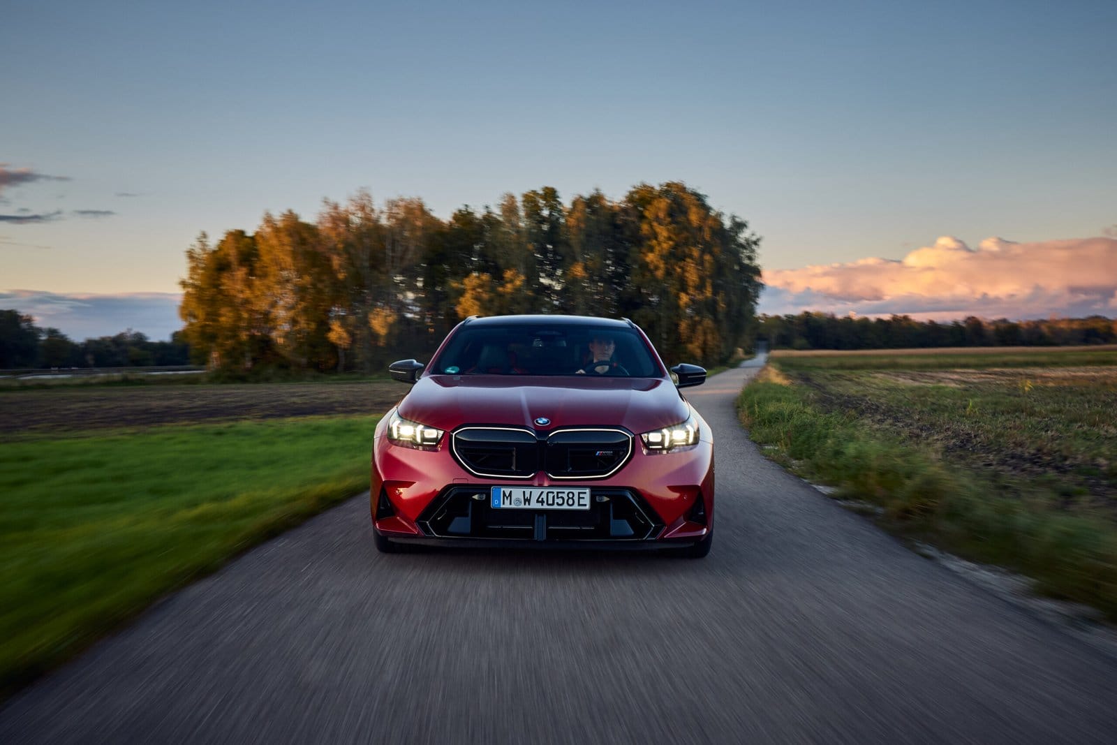 Red 2025 BMW M5 Touring Plug-in Hybrid driving towards camera on a rural road at sunset