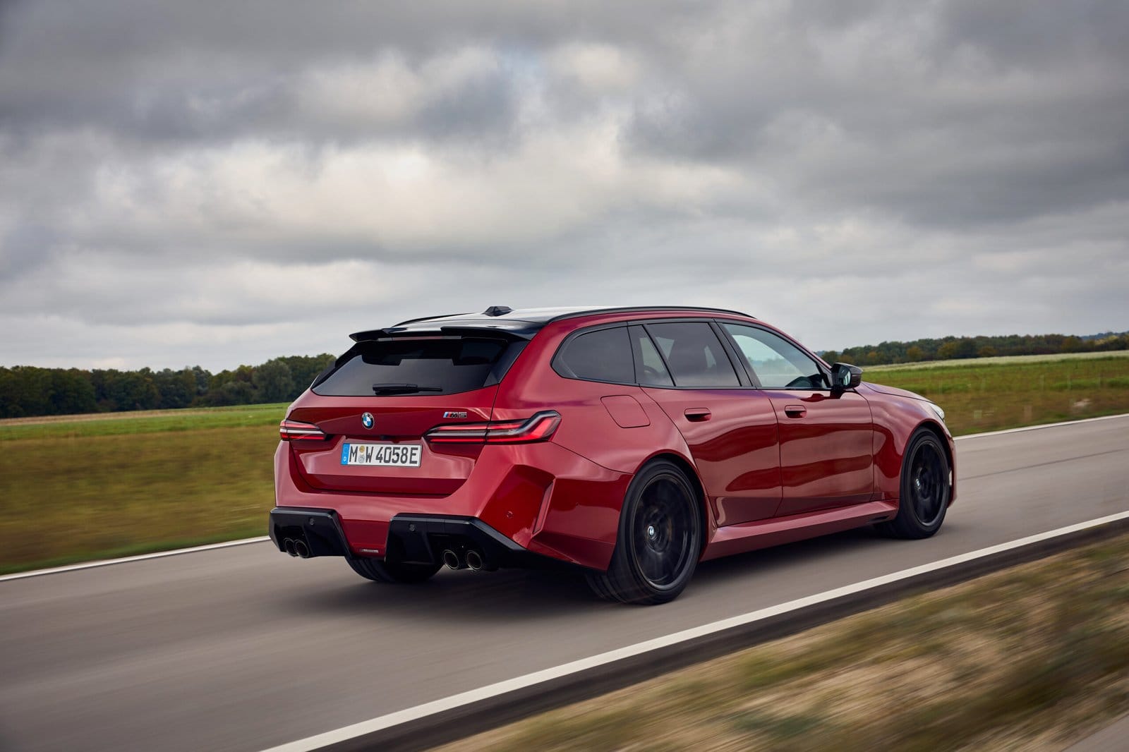 Red 2025 BMW M5 Touring Plug-in Hybrid driving on countryside road, viewed from rear side