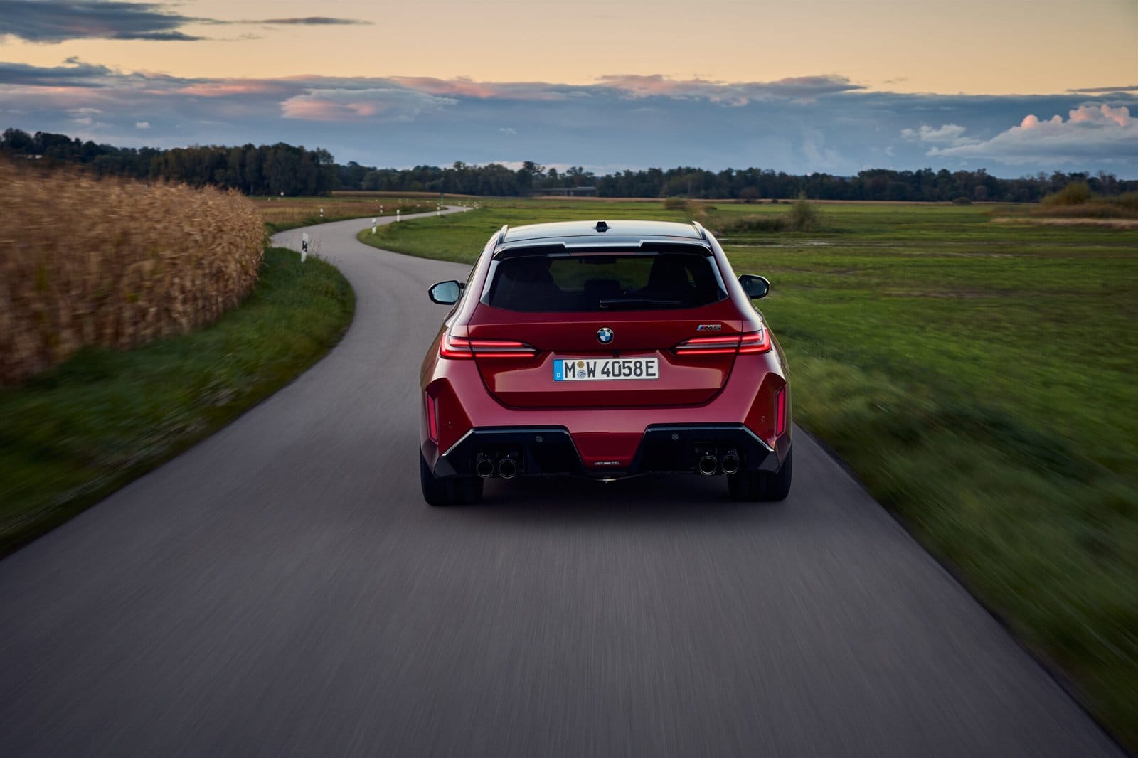 Rear view of red 2025 BMW M5 Touring Plug-in Hybrid driving through a curved road during sunset