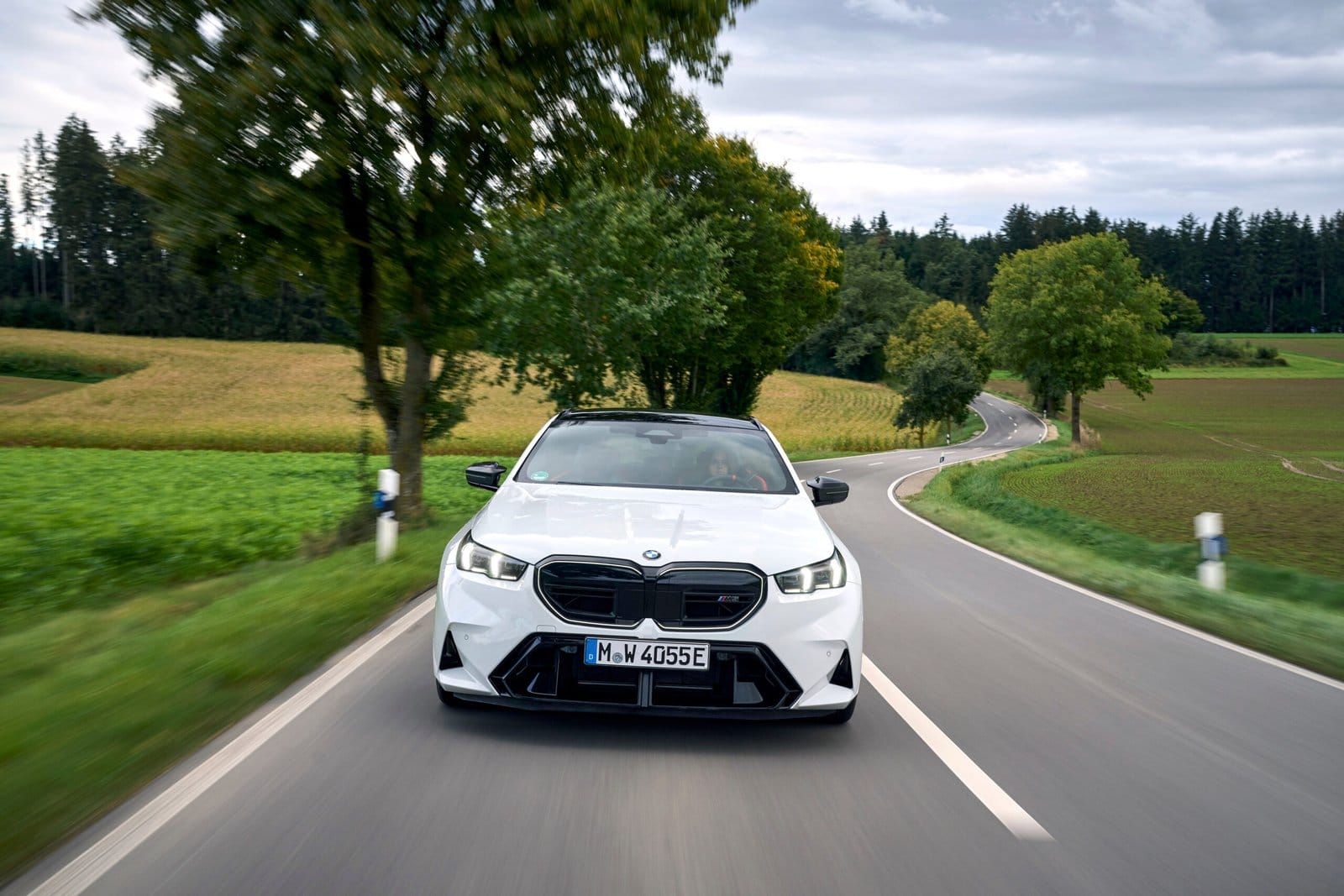 White 2025 BMW M5 Touring Plug-in Hybrid driving on winding rural road surrounded by trees and fields