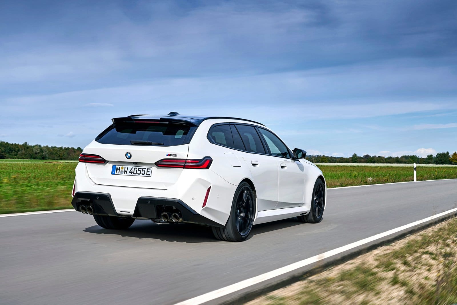 Rear-side view of 2025 BMW M5 Touring Plug-in Hybrid in white driving on a countryside road