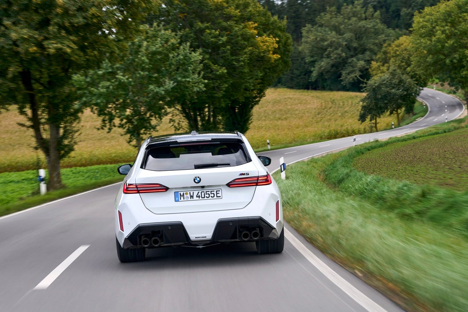 White 2025 BMW M5 Touring Plug-in Hybrid seen from behind driving through winding rural road with green landscape