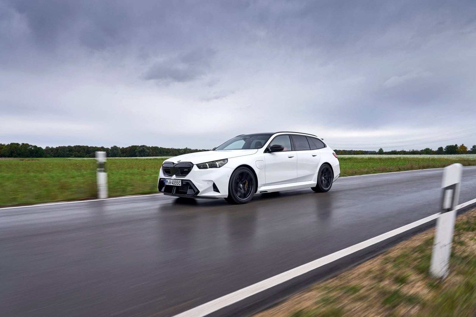 White BMW M5 Touring Plug-in Hybrid 2025 seen from the side front driving on a wet road with green fields and cloudy sky