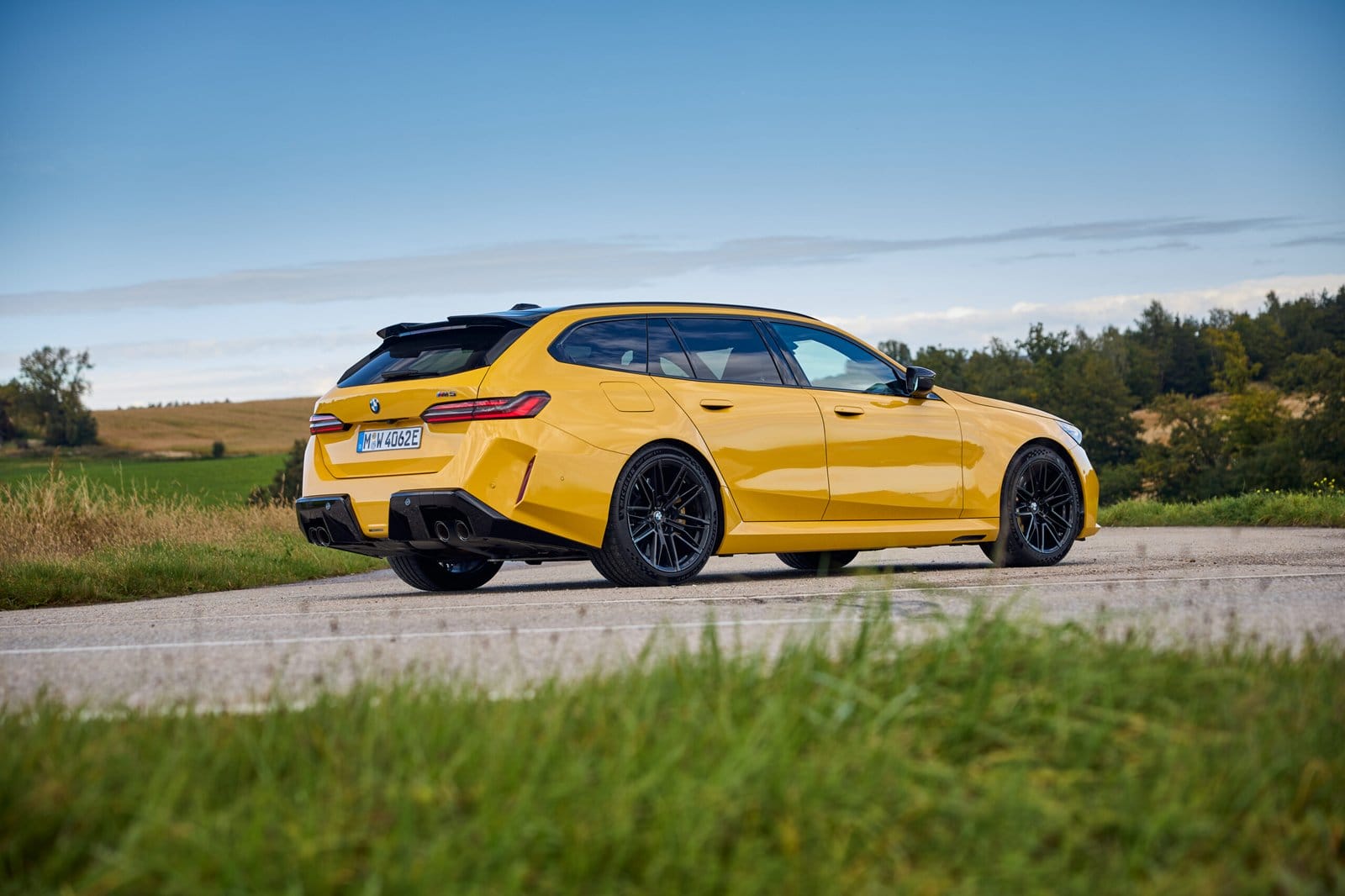 Rear-side view of the yellow 2025 BMW M5 Touring Plug-in Hybrid parked on a countryside road
