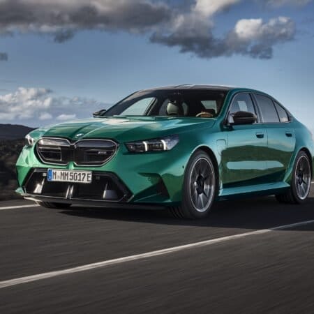 Front angle view of 2025 BMW M5 PHEV vehicle in green driving on a road, showing headlights, grille, and aerodynamic front design