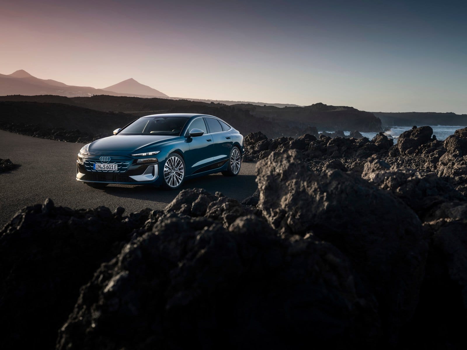Front view of Audi S6 e-tron electric vehicle parked on a road near rocky terrain and ocean coast at sunset.