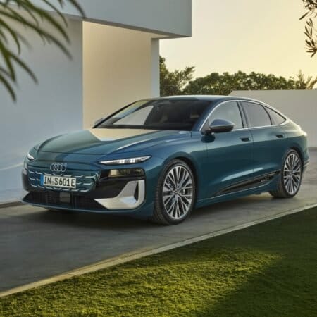 Front view of Audi S6 e-tron electric vehicle parked on a driveway near a modern white building and greenery.