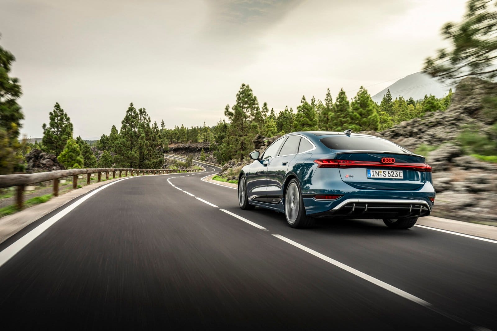 Rear view of Audi S6 e-tron electric vehicle driving on a winding forest road with trees and mountains in the background.