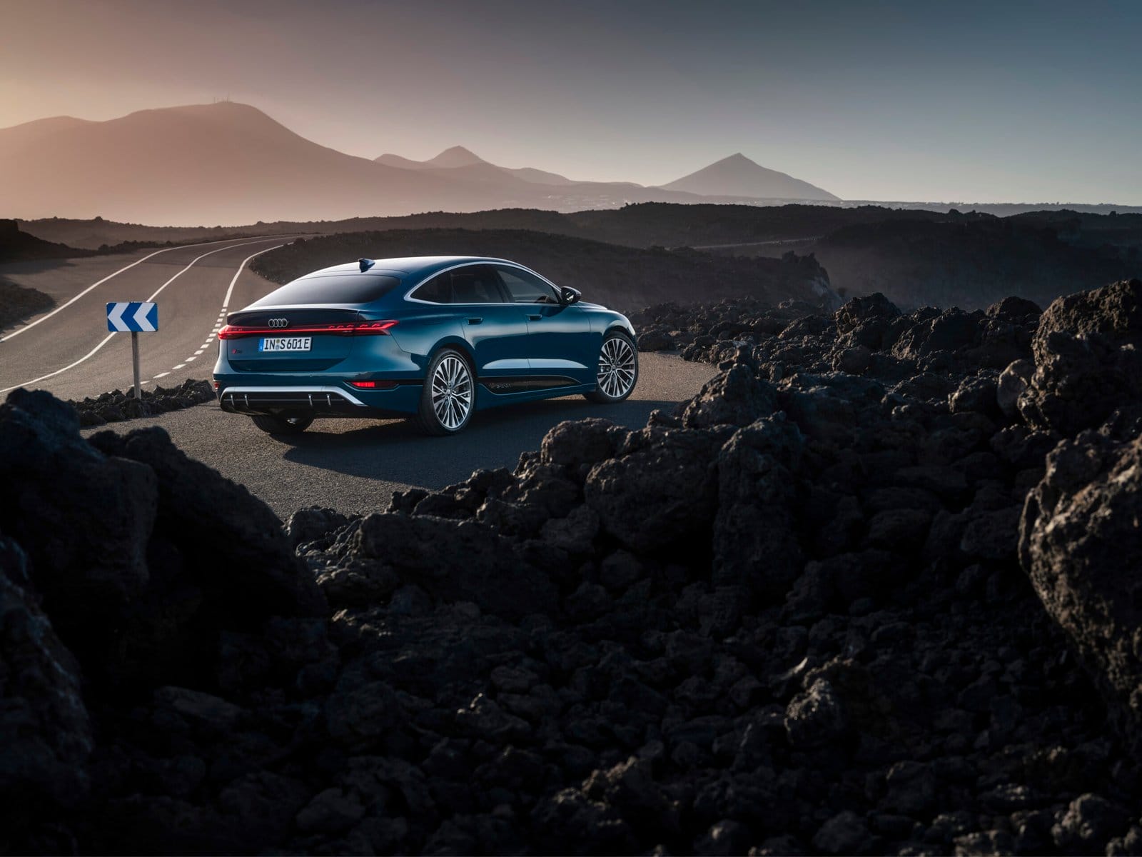 Rear view of Audi S6 e-tron electric vehicle driving along a winding road through volcanic terrain at sunset.