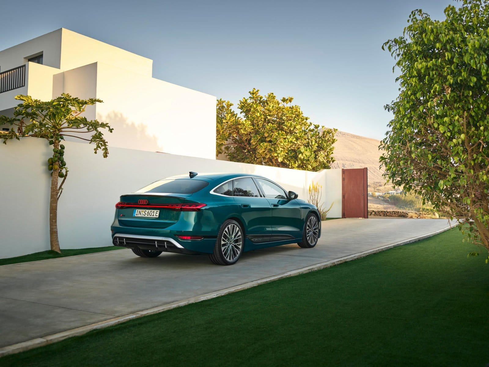 Rear side view of Audi S6 e-tron electric vehicle parked on a driveway beside a contemporary residential building and trees