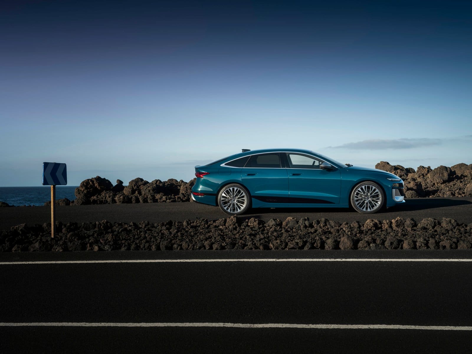 Side profile view of Audi S6 e-tron electric vehicle parked on a road near rocky coastal terrain under clear sky.