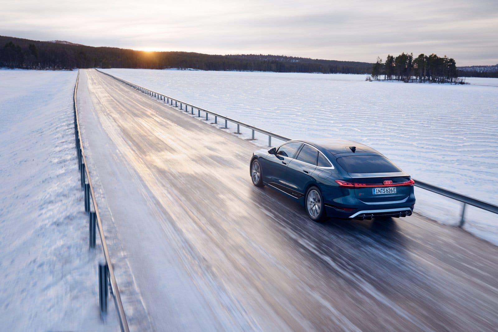 Audi S6 e-tron electric vehicle driving on a snow-covered road at sunset with forest and frozen lake in the background.