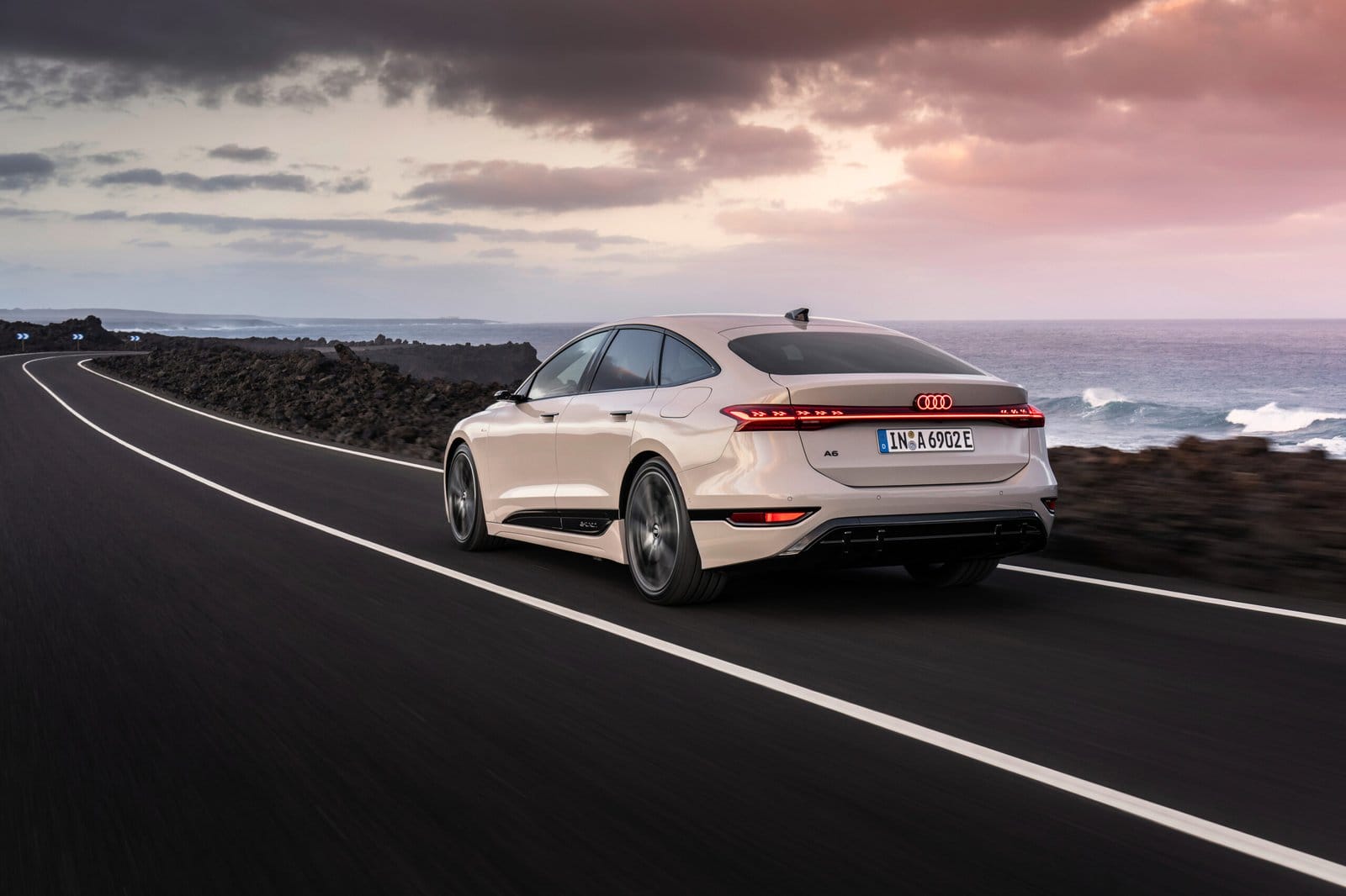 Rear view of the 2025 Audi A6 e-tron electric vehicle driving along a coastal road at sunset