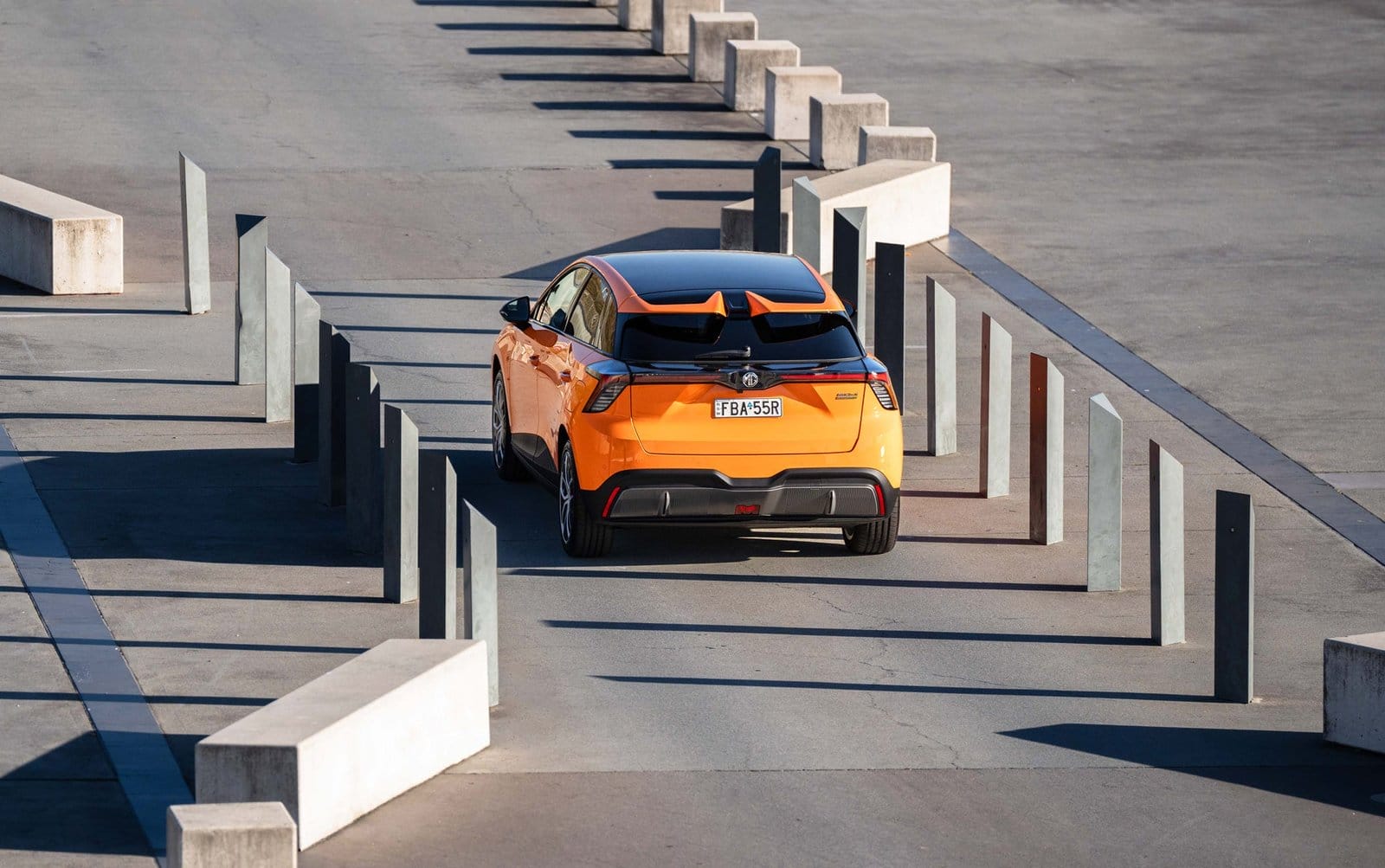 Rear view of the MG MG4 Electric Chinese EV driving through an urban space with geometric barriers and strong sunlight.